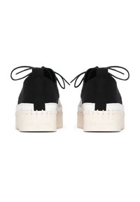 Zapatillas de lona negras con entresuela blanca, suela de goma y detalles decorativos de borlas. Cuentan con cordones y una textura suave en la parte superior.