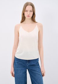 Beige spaghetti strap top with a V-neck design, made of a smooth, lightweight fabric. Paired with blue wide-leg jeans.