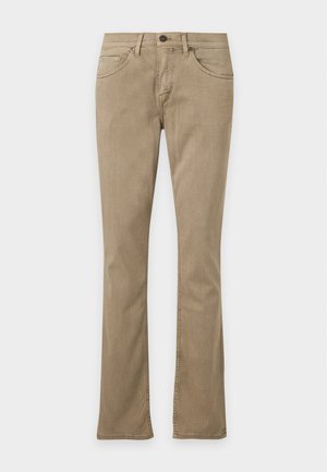 Homme portant une chemise à manches courtes à carreaux beige par-dessus un t-shirt blanc, un pantalon kaki et des baskets marron, debout devant un fond uni.