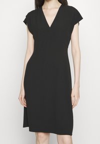 Svart klänning med V-ringning, korta ärmar och en figurnära silhuett. Tillverkad av ett slätt, matt tyg utan synliga mönster.