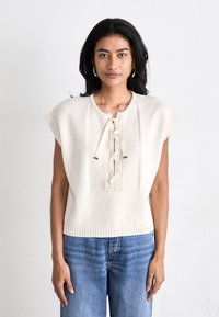 Knit cream-colored top with short sleeves, featuring a central lace-up detail and a ribbed hem. Worn with blue denim jeans.