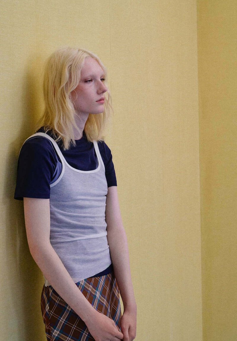 Jeune personne aux cheveux blonds mi-longs se tient contre un mur jaune pâle, portant un débardeur superposé et une jupe à carreaux.