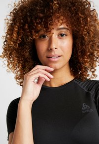 Black athletic shirt with short sleeves and textured shoulder accents. Features Odlo logo on the chest. Smooth, fitted design.