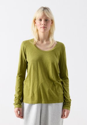 Lyshåret person iført en grøn langærmet top og lysegrå bukser, stående fremadrettet mod en ensartet hvid baggrund.