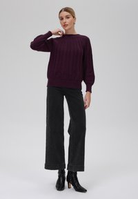 Strickpullover in tiefem Lila mit strukturiertem Muster auf der Vorderseite, geripptem Rundhalsausschnitt und überschnittenen Schultern; kombiniert mit hoch taillierten schwarzen weit geschnittenen Jeans und Stiefeln.