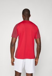 T-shirt de sport rouge à manches courtes, avec des accents rouges plus foncés et un logo doré dans le dos, fabriqué en tissu léger. Shorts blancs.