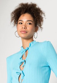 Top cano azul claro com nervuras, apresentando um decote alto com laços e uma abertura em forma de chave, acentuado por um padrão texturizado e mangas longas.