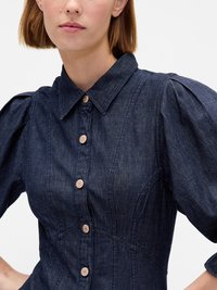 Abito in denim scuro con scollatura a collare, maniche a sbuffo corte e sette chiusure a bottoni con dettagli in legno circolare.
