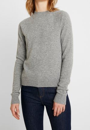 Femme portant un pull ajusté à manches longues gris clair et un jean bleu foncé sur un fond blanc uni.