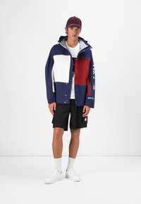 Farvet opdelt jakke i blå, hvid og rød; sat sammen med sorte shorts. Har hætte, knapper og branddetaljer. Hvide sneakers og sokker.