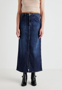 Denim maxi skirt features a dark wash, front slit, two large front pockets, and contrast stitching. Paired with black ankle boots.