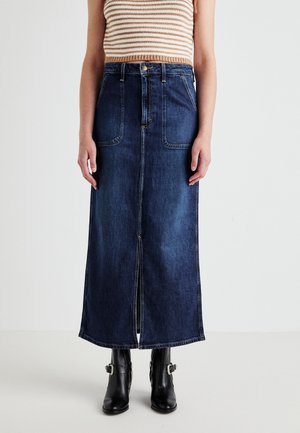 Denim maxi skirt features a dark wash, front slit, two large front pockets, and contrast stitching. Paired with black ankle boots.