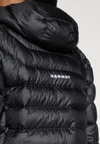 Černá zateplená péřová bunda s kapucí, s prošívanými částmi a jemným mřížkovaným vzorem. Logo "MAMMUT" bílé barvy na zadní straně.