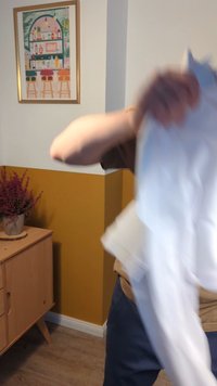 Vit skjorta i rörelse, tillverkad av tyg, med en mjuk textur. I bakgrunden syns en gyllene vägg och en träbyrå med blommor.
