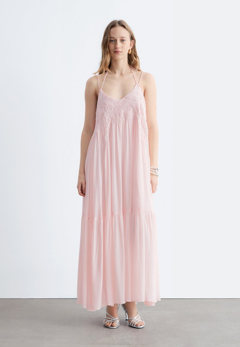 Jeune femme debout portant une robe maxi fluide rose pâle sans manches avec des détails en dentelle et des talons argentés à brides sur un sol gris.