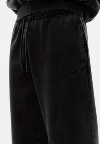 Čierne tepláky s elastickým pásom a šnúrkou na stiahnutie. Mäkký materiál, bočné vrecká a vyšívaný logo detail na stehne.