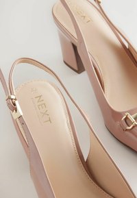 Pantofi cu toc înalt de culoare roz pal, cu un design tip slingback, având o cataramă aurie și brandingul "NEXT Forever Comfort" pe tălpă.