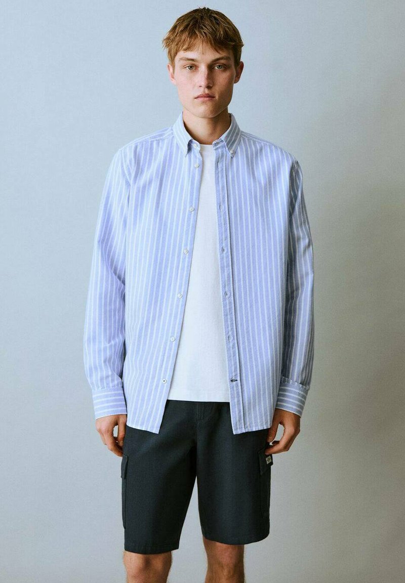 Un giovane uomo è in piedi contro uno sfondo grigio chiaro, indossando una camicia a righe blu sopra una t-shirt bianca e pantaloni cargo scuri. Espressione seria.