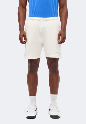 Άτομο που φορά λευκά σορτς με κορδόνι, λευκά αθλητικά παπούτσια, λευκές κάλτσες και μπλε πουκάμισο, στέκεται με τα χέρια χαλαρά στο πλάι.