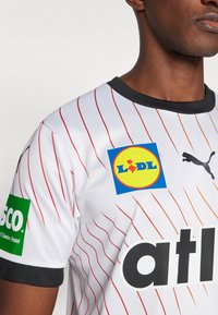 Bijela sportska majica s crvenim dijagonalnim prugama, crnim ovratnikom, koja sadrži Puma logo i sponzorske naljepnice u plavoj i zelenoj boji. Lagani materijal.