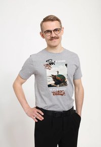 Šedé tričko s krátkým rukávem s grafickým potiskem, který zahrnuje text ve graffiti stylu a obrázek skateboardisty na barevném, ošumělém objektu.