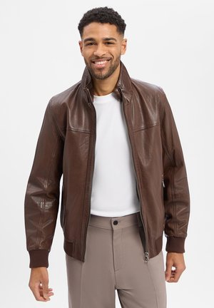 Lächelnder Mann mit brauner Lederjacke mit Reißverschluss, weißem Hemd und beiger Hose vor hellem, einfarbigem Hintergrund.