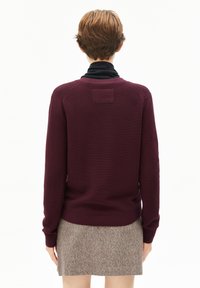 Persoon met kort bruin haar, gekleed in een donkerrode trui over een zwart shirt en een grijze rok, met de rug naar de camera tegen een witte achtergrond.