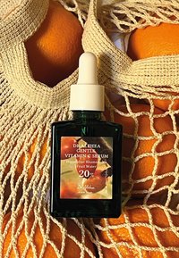 Botella de vidrio verde con un gotero blanco, que presenta una etiqueta colorida. Rodeada de naranjas y una bolsa de malla beige.