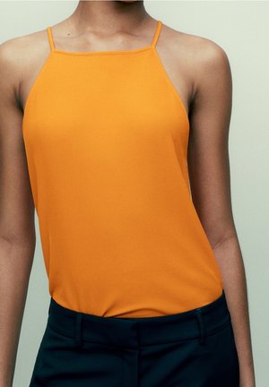 Kvinde iført ærmeløs orange haltertop stoppet ned i sorte bukser foran en ensartet lys baggrund.