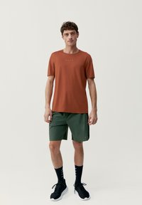 Pánske tričko v rustikálnej oranžovej farbe, s okrúhlym výstrihom a krátkymi rukávmi; skombinované so zelenými šortkami športového strihu. Čierne ponožky a bežecké tenisky dopĺňajú vzhľad.