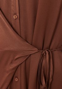 Gros plan d'une robe en tissu marron avec des boutons et une ceinture nouée à la taille, montrant des détails de texture et de couture.