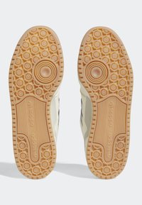 Suelas de zapatillas de goma con un agarre texturizado. Color beige claro con un patrón circular y la marca Adidas en el lateral.
