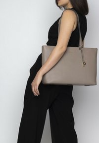 Tote bag in licht taupe leer met een gladde textuur, minimalistisch ontwerp, ritssluiting en een verstelbare strap. Gouden logo accent op de voorkant.