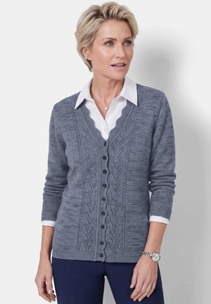 Vrouw met kort blond haar draagt een blauw gemêleerde gebuttonde cardigan over een wit overhemd met kraag en een donkere broek, kijkt opzij.