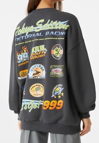 Sudadera negra de gran tamaño con gráficos y texto coloridos relacionados con las carreras de motocicletas, con varios parches y un cuello acanalado.