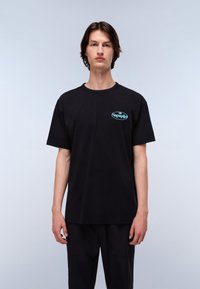 Sort bomulds t-shirt med rund halsudskæring, korte ærmer og et teal logo på venstre bryst. Modellen står foran en lysegrå baggrund.