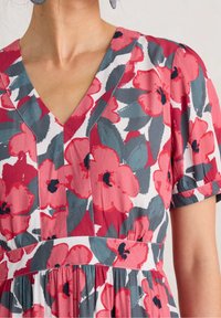 Kvinna iklädd en kortärmad klänning med röda och rosa blommönster samt gröna löv på vit bakgrund, med en V-ringning.