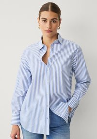Camisa de botones a rayas azules y blancas con cuello, de mangas largas y corte relajado. Cuenta con un bolsillo en el lado izquierdo.