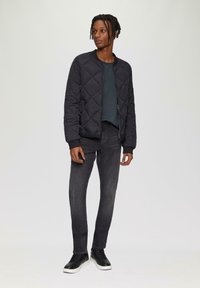 Zwarte gewatteerde bomberjack met geribde manchetten, gedragen over een donkergroen topje, gecombineerd met slim-fit zwarte jeans en zwarte sneakers.