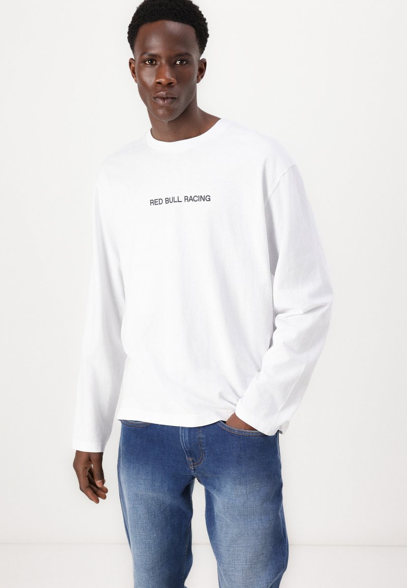 Weißes Langarm-T-Shirt mit "RED BULL RACING" in Schwarz auf der Vorder- und Rückseite, kombiniert mit blauen Denim-Jeans.