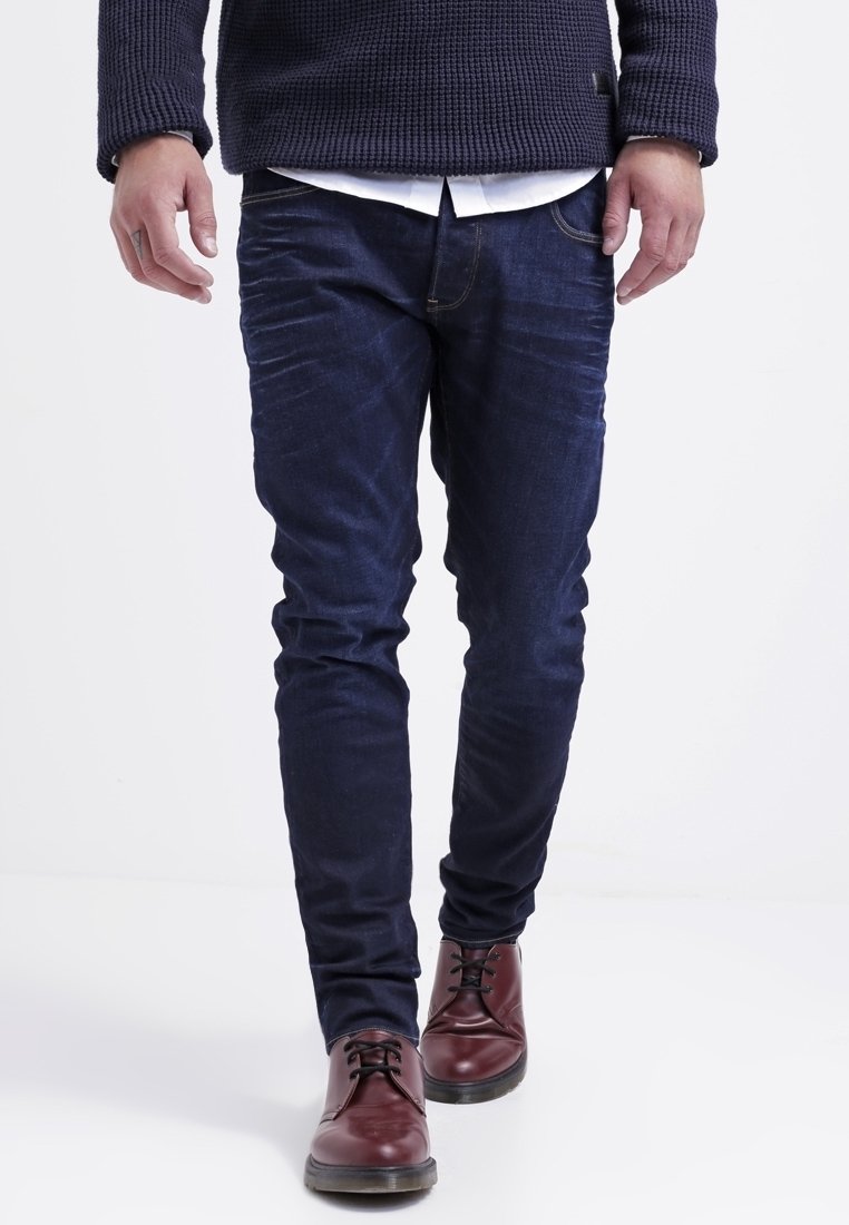 Jeans blu scuro in perfetto slim fit con leggeri effetti di strappo, abbinati a scarpe di pelle marrone con lacci. Maglione in maglia con tessuto testurizzato sopra.