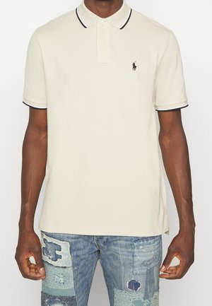 Mężczyzna w białej koszulce polo z krótkim rękawem z czarnym obszyciem i haftowanym logo, w niebieskich dżinsach typu patchwork.