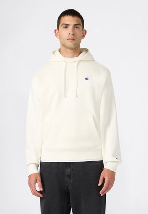 Mann, der nach vorne schaut, trägt einen elfenbeinfarbenen Hoodie mit Kordeln, einem kleinen Logo auf der Brust und am Ärmel sowie schwarze Hose.