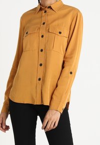 Kvinna iförd en senapsgul skjorta med knappar och två bröstfickor, klädd i svarta byxor, stående mot en enkel vit bakgrund.