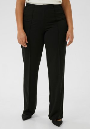 Femme portant un pantalon noir taille haute à jambes droites et des chaussures noires pointues, debout devant un fond blanc uni.