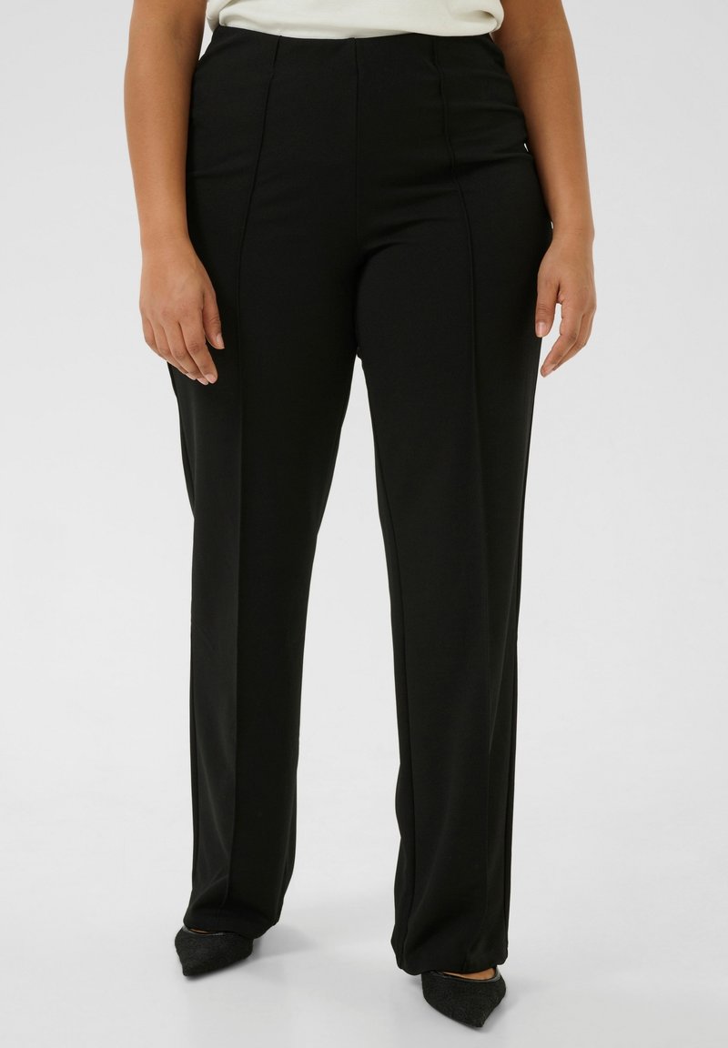 Femme portant un pantalon noir taille haute à jambes droites et des chaussures noires pointues, debout devant un fond blanc uni.
