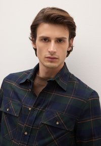 Camisa de franela para hombre, con patrón de cuadros en verde oscuro y azul marino, dos bolsillos en el pecho con cierre de botones, cuello abotonado, textura suave.