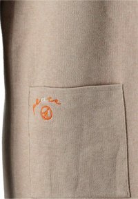 Beige tygficka med det lilla orange broderade ordet "peace" och en fredssymbol nära det övre vänstra hörnet av fickan.