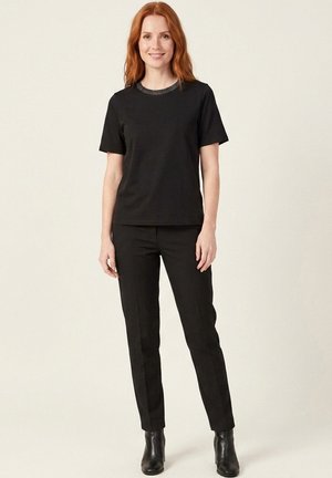 Femme rousse debout, portant un haut noir à manches courtes avec un col à motifs, un pantalon noir ajusté et des bottines noires, souriante.