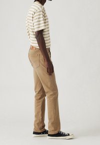 Pantalon en coton beige à jambes droites, avec poches latérales et poches plaquées à l'arrière. Associé à une chemise à manches courtes rayée et des baskets noires.